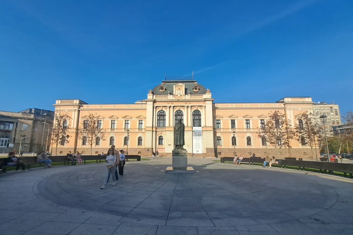 Popločan trg u centru Kragujevca sa spomenikom ispred velike žute zgrade iz 19. veka pod vedrim nebom