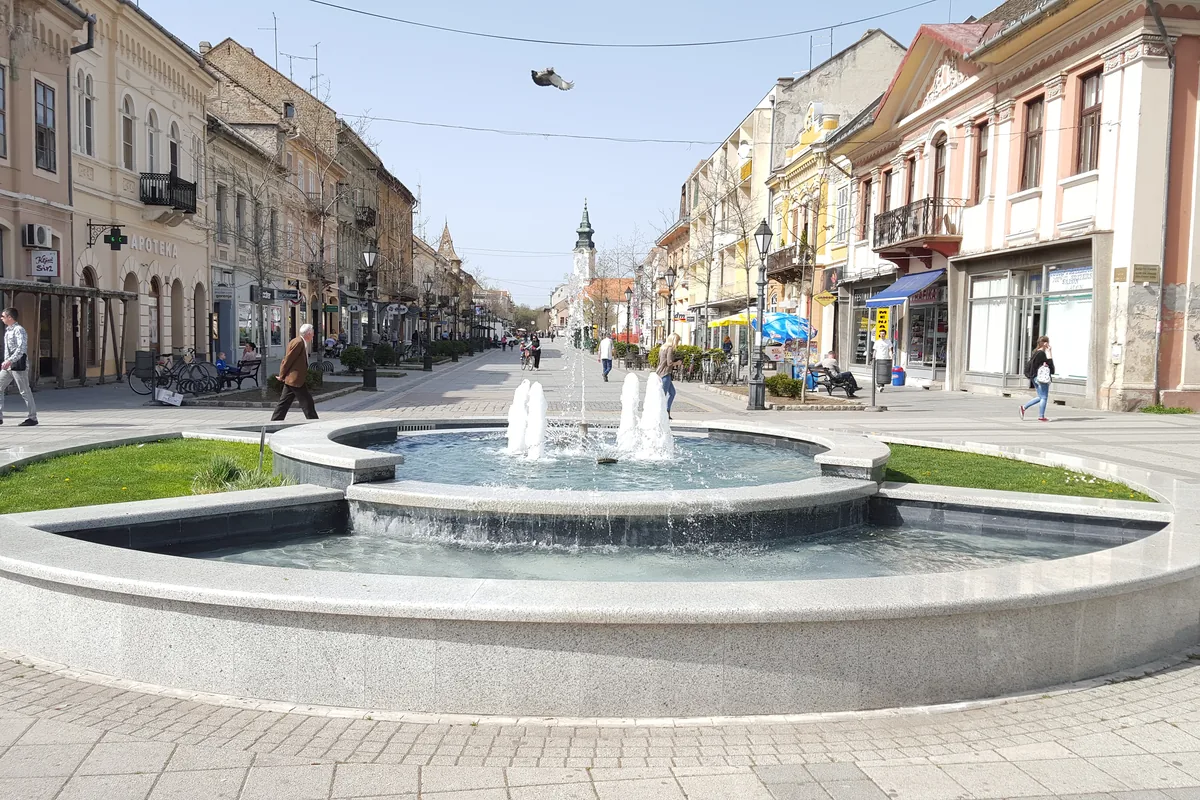 Pešačka glavna ulica u Somboru sa fontanom u prvom planu, nizom starih zgrada i zvonikom u daljini