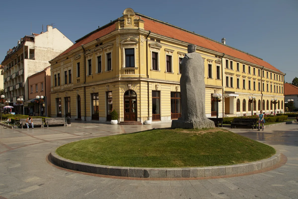 Pešačka ulica u Valjevu, žuta zgrada iz 19. veka i kameni spomenik na travnatom ostrvu