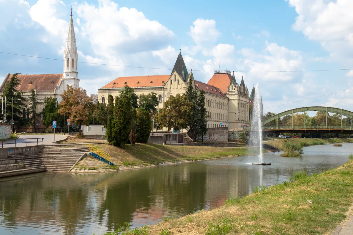 Palata pravde i vitki zvonik Reformatske crkve u Zrenjaninu iznad vode sa fontanom, lučni gvozdeni most u pozadini