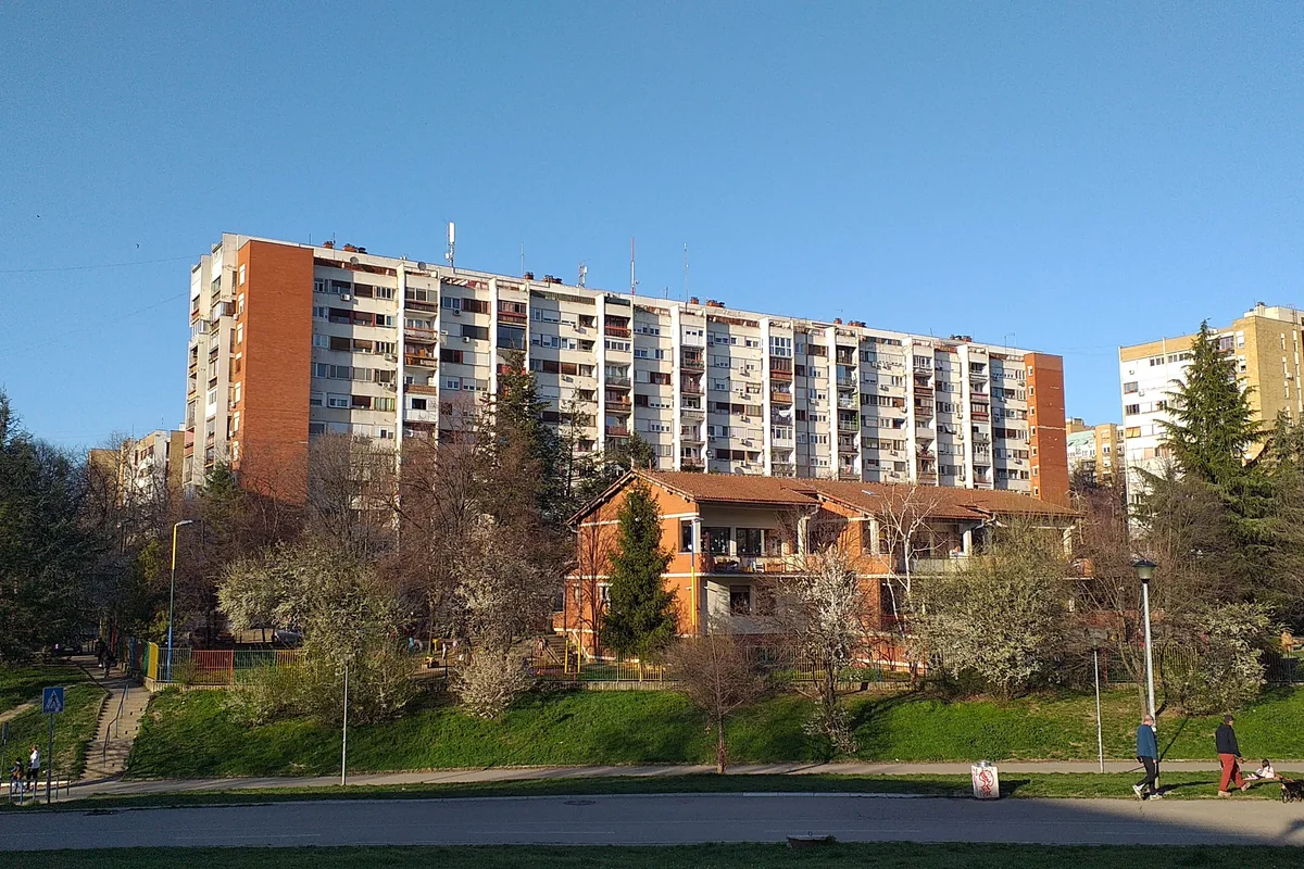 Visoka stambena zgrada i niža zgrada vrtića okružene drvećem u Mirijevu II, sunčan dan